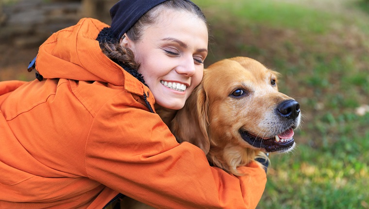 Smiling young woman kneeling and embracing a dog. Wears casual clothes, with eyes closed. Focus on foreground.