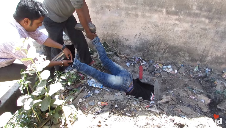 Two men lower another by his feet into the septic tank.