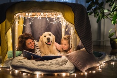 Young happy mother reading a book to her small girl while being in a tent at home with their dog.