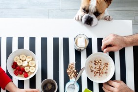 Tavolo della cucina con commensali. Ciotole di porridge di avena e frutta fresca. Mani che impugnano tazzine di caffè espresso e ciotole di ceramica colme di banana a fette, lamponi, avena e mandorle. Bicchiere di vetro con latte e spuma. Caffè macchiato.