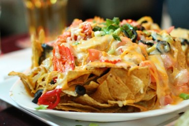 Delicious heaping plate of nachos and cheese with olives and peppers. Beer in background.