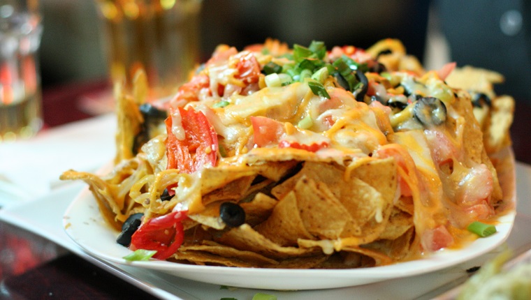 Delicious heaping plate of nachos and cheese with olives and peppers. Beer in background.