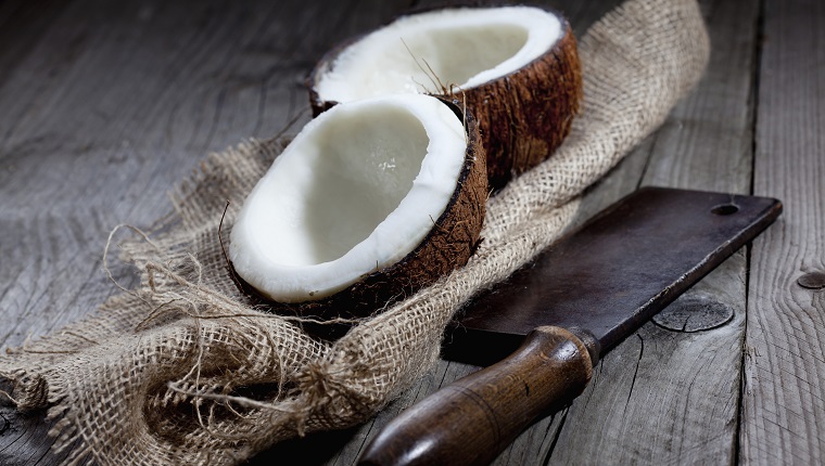 Kokosnuss (Cocos nucifera) auf Sackleinen, altes Hackmesser,grauer Holzuntergrund, Holztisch