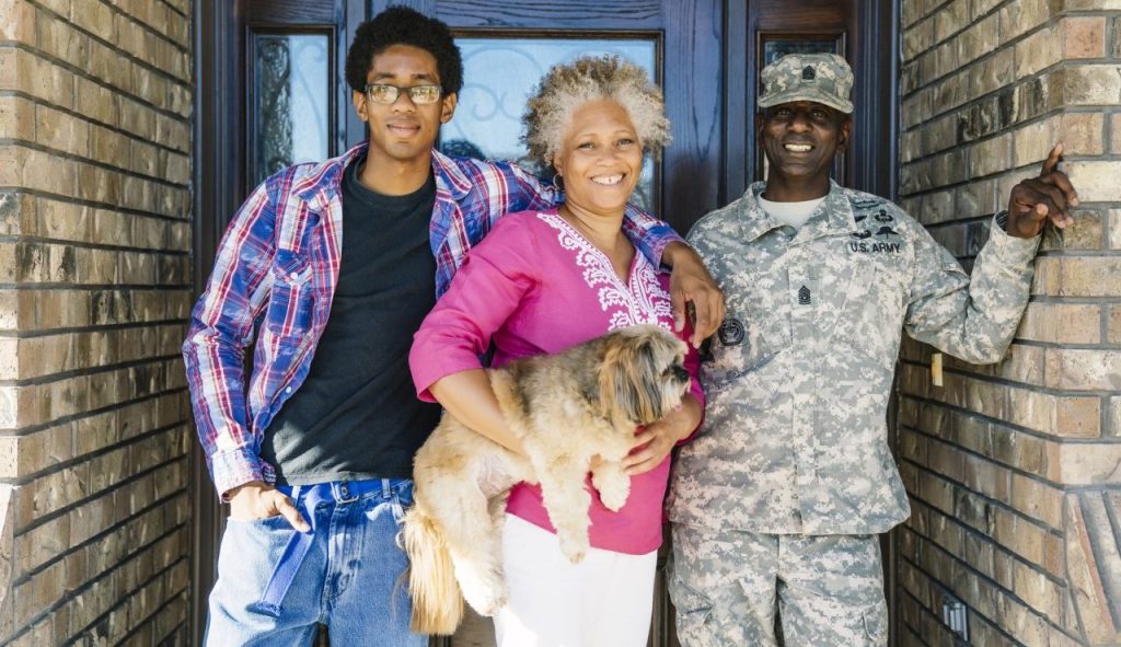 Black family of 3 members-including a son, mom, and father in a Armed Forces uniform-standing in doorway with mom holding their small dog.