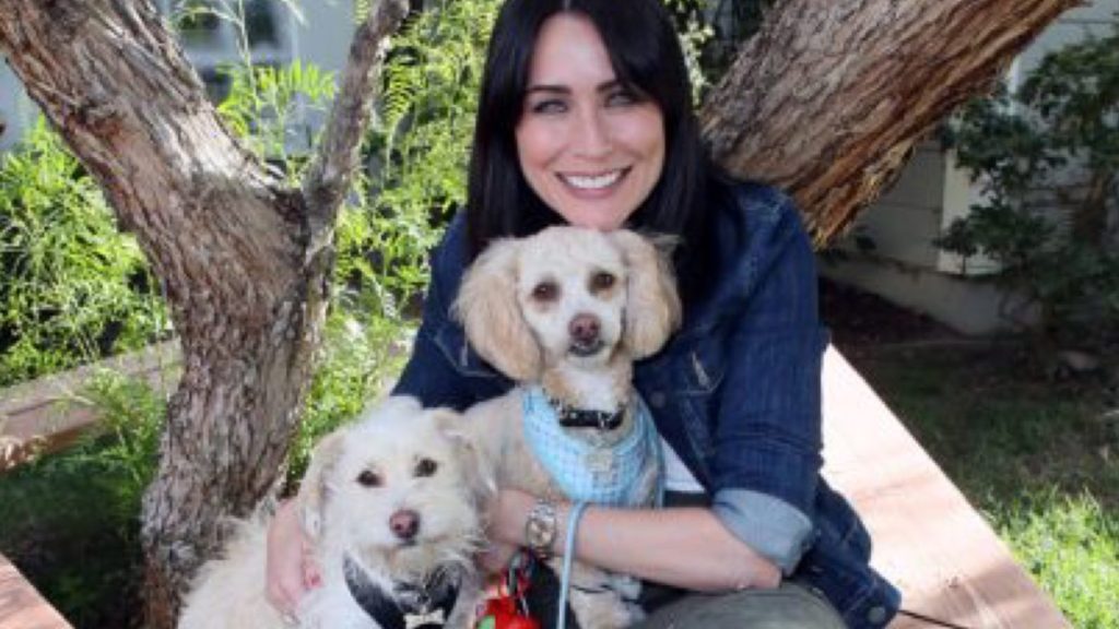 Actress Rena Sofer sitting on a bench holding two small white dogs, Rena Sofer's dog, Alfie,has died of cancer