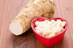 Horseradish root and shredded horseradish in a heart-shaped bowl on a wooden surface.